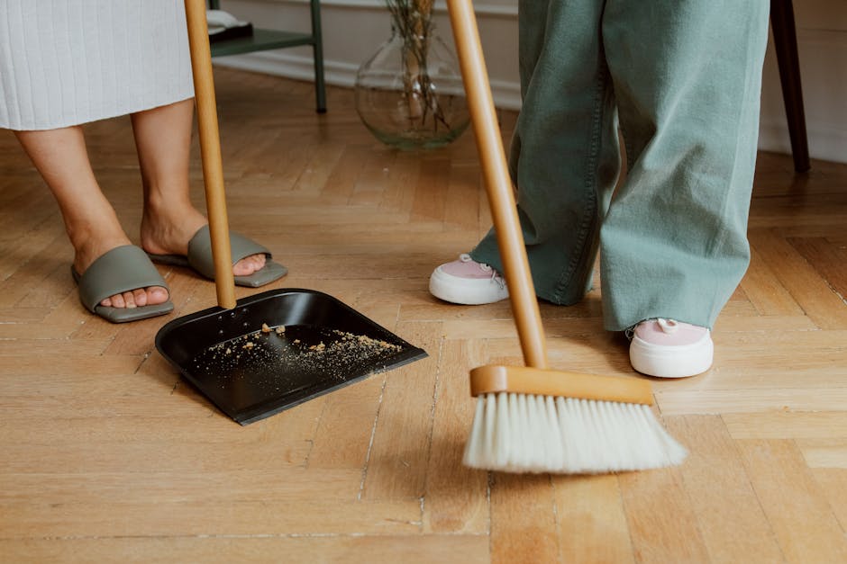 Close-up of a domestic cleaning scene showing the lower legs and feet of two individuals, one wearing grey slide slippers and light-colored pants, the other in white sneakers and green pants, standing on a wooden floor. A dustpan containing debris is positioned on the floor, with a broom resting against the dustpan, indicating surface cleaning. The background includes a part of a room with warm lighting, a glass vase, and minimal furniture, reflecting a tidy, well-maintained indoor space. The scene illustrates routine surface cleaning and debris removal, consistent with professional or household hygienic practices. Cleaners Kentish Town utilizes thorough cleaning methods to ensure surface sanitisation and dust-free environments.