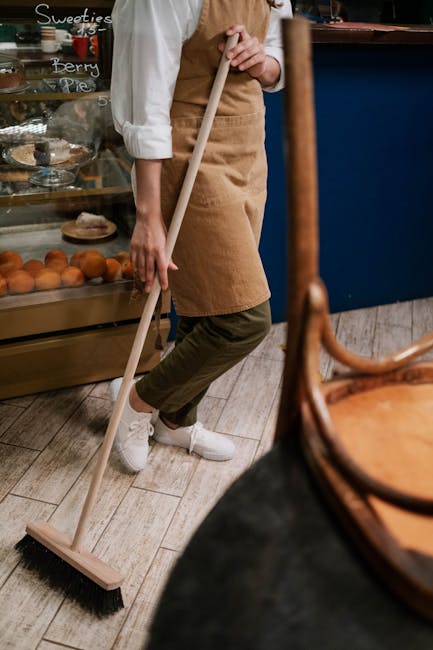 A person wearing a white shirt, beige apron, and khaki pants is using a broom to sweep the wooden floor of a bakery or café. The background shows a glass display case with baked goods like bread and pastries, and handwritten chalkboard signs advertising sweet treats such as berry pie. The space is well-lit, with natural light highlighting the cleanliness of the surface, and the floor appears dust-free and polished. The individual is focused on maintaining hygiene and cleanliness, exemplifying domestic or commercial surface cleaning, which Cleaners Kentish Town offers to avoid hidden charges in Kentish Town.