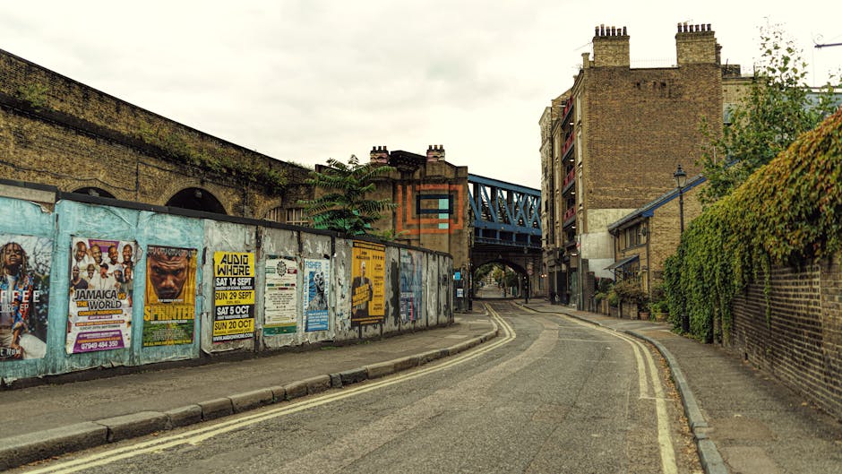 A narrow urban street in Kentish Town featuring residential buildings and a railway bridge overhead. The road surface is asphalt with visible double yellow lines along the curb. On the left side, a weathered metal fence displays various colorful posters and flyers, some slightly torn or faded. The buildings on the right are brick with some greenery climbing the walls, and the street is illuminated by natural daylight under an overcast sky. No visible cleaning activity or tools are present in this image, but it reflects typical cityscape elements associated with access challenges in the area. Cleaners Kentish Town offers professional cleaning services to maintain hygiene and order in such environments.