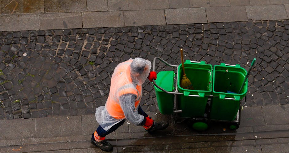 A commercial cleaner from Cleaners Kentish Town dressed in a high-visibility orange vest, a grey top, and red gloves, is pushing a green cleaning cart on a wet cobblestone street in Kentish Town. The cart contains multiple green bins, one of which holds a yellow mop with a wooden handle. The surrounding surface includes a concrete sidewalk and an area of textured cobblestones, both appearing damp, indicating recent or ongoing cleaning activities. Natural daylight illuminates the scene, highlighting the cleanliness of the surfaces. This image exemplifies surface cleaning and maintenance performed by professional cleaners to ensure hygiene and cleanliness in public urban environments, aligning with the services offered by Cleaners Kentish Town for both residential and commercial clients.