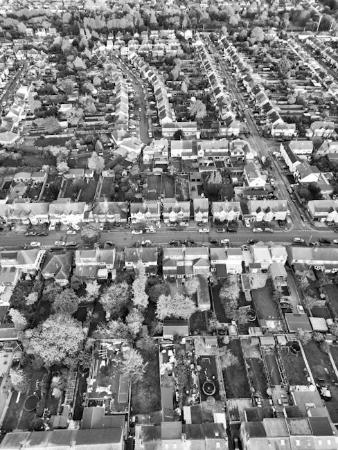 An aerial view in black and white showing a residential area with rows of terraced houses and detached homes, featuring rooftops, backyards, and garden spaces. The streets are lined with trees and vehicles, with visible sidewalks. The scene includes various surfaces such as tiled and paved roofs, smooth asphalt roads, and landscaped garden areas, some with visible outdoor furniture, fences, and lawn patches. The lighting highlights the clean, well-maintained appearance of the urban environment. This image exemplifies the type of detailed surface cleaning and maintenance services offered by Cleaners Kentish Town in the Kentish Town area, focusing on residential deep cleaning and hygiene upkeep.