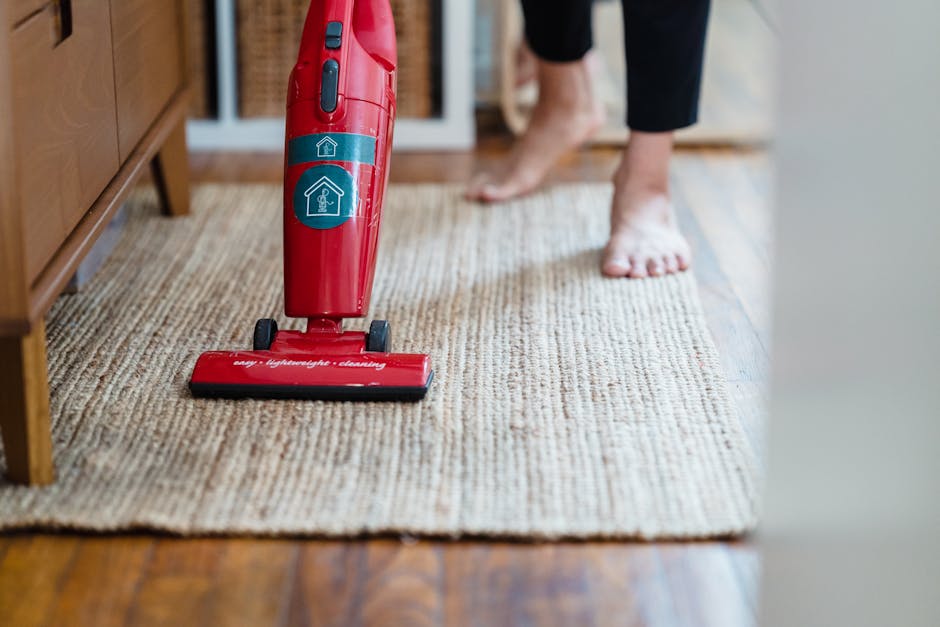 Close-up of a vacuum cleaner head placed on a patterned, beige carpet in a residential room. The vacuum is black with a slightly textured surface, and a visible control button on top. The carpet featu