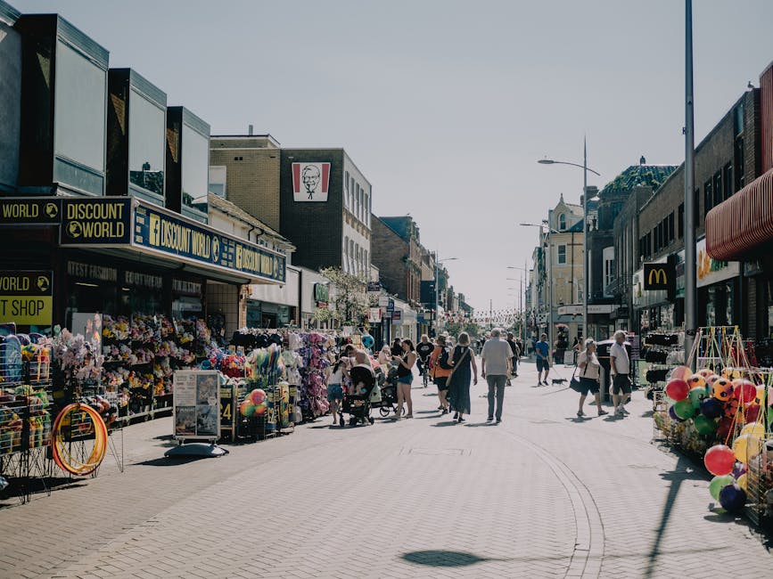 A busy pedestrian street in a commercial area with various shops and stalls, including a souvenir and balloon stall on the left, and storefronts like discount shops and fast-food outlets, visible signs such as 'DISCOUNT WORLD' and a McDonald's. The pavement is clean and well-maintained, with a mix of people walking, shopping, and pushing strollers. The buildings are multi-story with large windows and signage, and the sky is clear and bright, indicating daytime. The scene reflects a lively, clean urban environment, captured for a residential or commercial cleaning service page by Cleaners Kentish Town, emphasizing surface cleaning and maintenance of public spaces. Ready for deep cleaning, hygiene, and sanitisation standards within a bustling town centre. The overall atmosphere is vibrant and orderly, showcasing a well-kept shopping street.