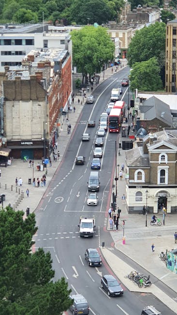 An aerial view of Fortess Road in Kentish Town, showing a busy street with parked and moving cars, including a double-decker bus, alongside low-rise commercial and residential buildings with varied brick facades. The scene features sidewalks with pedestrians, some crossing at intersections, and is lined with green trees providing shade. The lighting indicates a clear day, and the overall scene appears tidy and well-maintained, reflecting a typical urban setting. This image visually represents the area where Cleaners Kentish Town offers surface cleaning, deep cleaning, and sanitisation services, highlighting the importance of maintaining cleanliness in busy residential and commercial streets.
