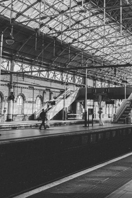 Inside a large, industrial-style train station with a high arched metal ceiling and exposed beams. The platform has multiple stairs leading to an upper level, with a few passengers waiting or walking. The floor appears clean and smooth, likely made of concrete or stone, and is well-maintained. The station is illuminated by natural light coming through large windows and skylights. Metal train tracks run along the platform edge, and electronic signage displays information. A person is seen walking across the platform, and another is standing near a support column. The overall scene highlights a spacious, hygienic environment typical of a busy transportation hub, with the work of professional cleaning services like Cleaners Kentish Town ensuring a pristine, well-maintained setting.
