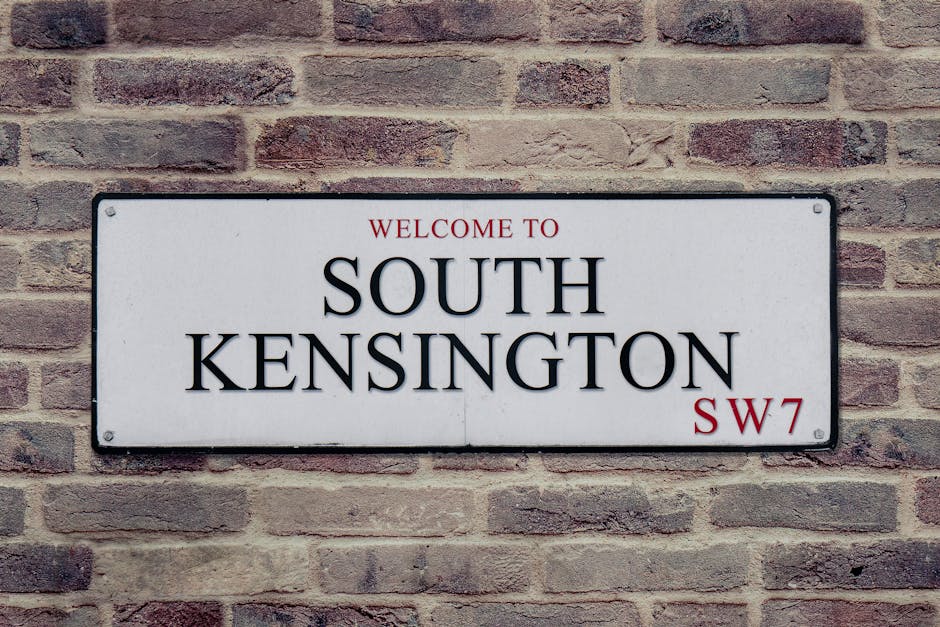 A rectangular white sign with black lettered text that reads 'Welcome to South Kensington SW7' mounted on a brick wall with reddish-brown bricks. The sign has a simple black border and is photographed in natural daylight, indicating a well-maintained exterior. This image illustrates the importance of surface cleaning and maintenance of signage for hygiene and presentation, consistent with professional cleaning standards promoted by Cleaners Kentish Town in relation to spotting poor cleaning workmanship. The bricks and sign are clean and free of dust or stains, highlighting good surface scrubbing and preservation.