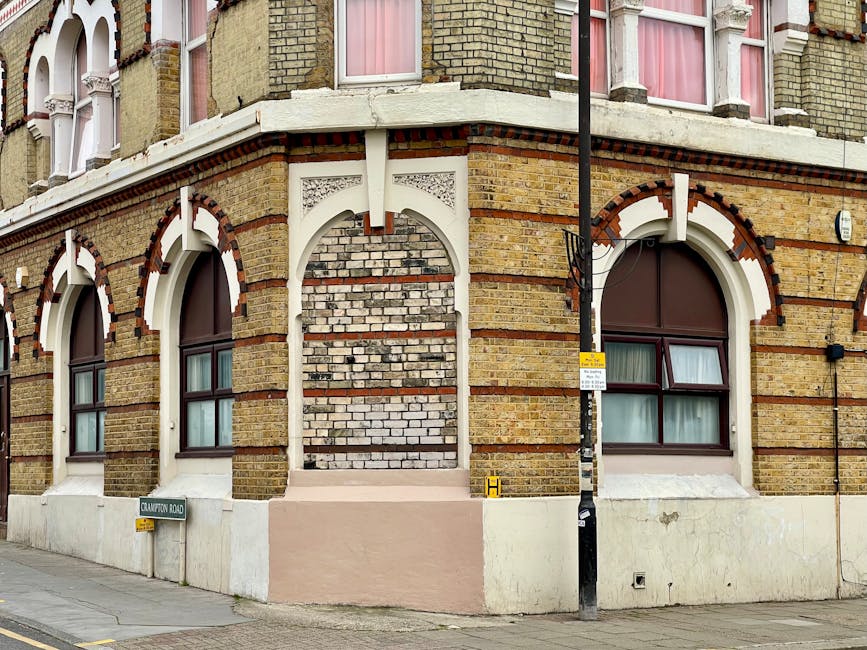 Close-up view of a historic brick corner building in Kentish Town showcasing a mix of beige, red, and white bricks with decorative arch window frames. The ground level features a smooth, white-painted surface with two large windows fitted with brown frames and partially obscured curtains. A street sign reading 'Crawton Road' is visible at the base, along with a black street lamp pole positioned in front. The building's exterior appears clean but exhibits some signs of weathering typical of aged brickwork, with no visible cleaning tools or products in the image. The scene is illuminated by natural daylight, highlighting the textures and colors of the brick surfaces, with a focus on the structure's architectural details. Cleaners Kentish Town would conduct surface cleaning and maintenance to preserve the building's appearance.
